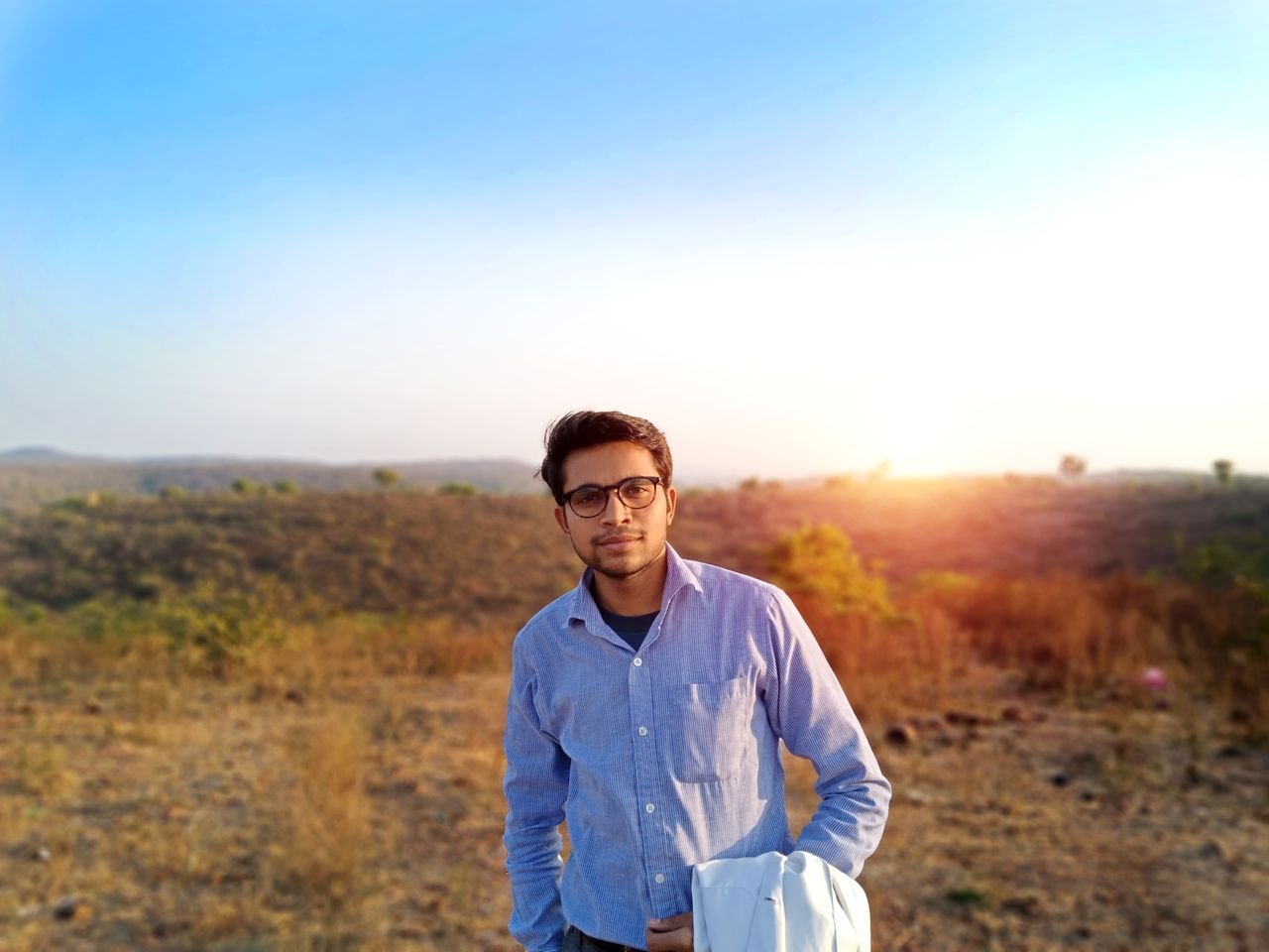 Portrait of a young man standing in an Indian field at sunset, dressed casually, with the sun setting in the background.